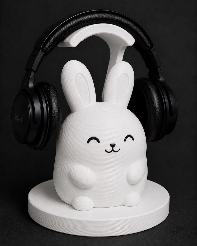 May include: A white bunny-shaped headphone stand with a smiling face and black accents. Black headphones rest on the stand. The stand is on a white circular base, all against a dark background.