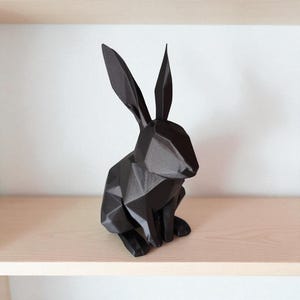 May include: A black geometric rabbit sculpture with long ears, sitting on a light-colored wooden shelf. The rabbit is made of multiple flat surfaces, giving it a modern, angular appearance. The background is a plain white wall.