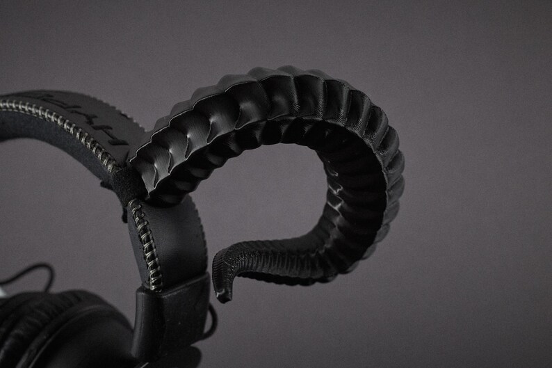 May include: Black headphones featuring a distinctive, curved horn-shaped design. The headphones have a padded headband and earcups, with visible stitching. The horn-like structure is textured, adding a unique visual element.