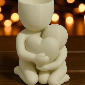 May include: A cream-colored sculpture of two figures embracing a heart. One figure kneels, cradling the heart, while the other stands above, forming a loving embrace. The sculpture is made of a smooth, matte material.