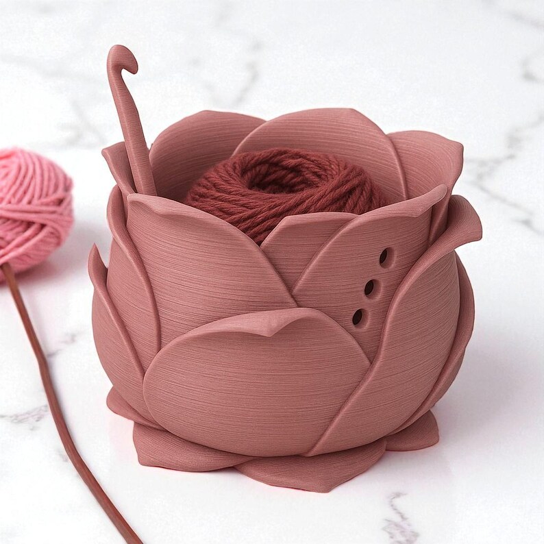 May include: A dusty rose-colored yarn holder shaped like a rose, with a crochet hook and a ball of yarn inside. The holder has three small holes on the side. A pink ball of yarn is on the left.