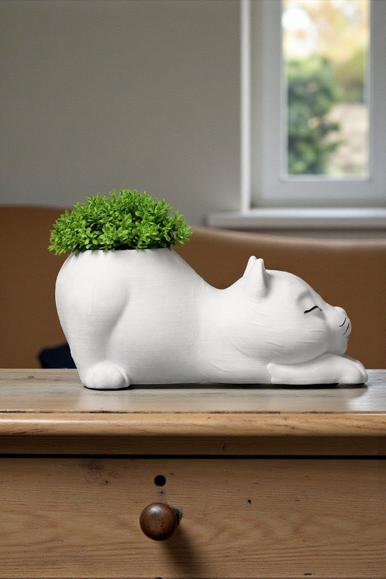 May include: A white ceramic cat-shaped planter with a green plant sprouting from its back. The cat is lying down with its eyes closed. The planter is on a wooden table.
