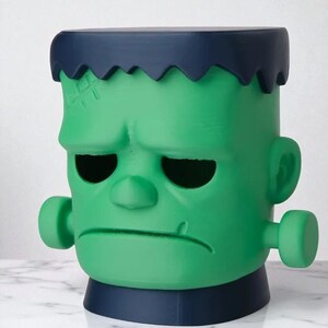 May include: A green Frankenstein-themed decorative container with a dark blue top and base. The container has a detailed face with stitched scars, black eye sockets, and side bolts. A unique Halloween or novelty item.