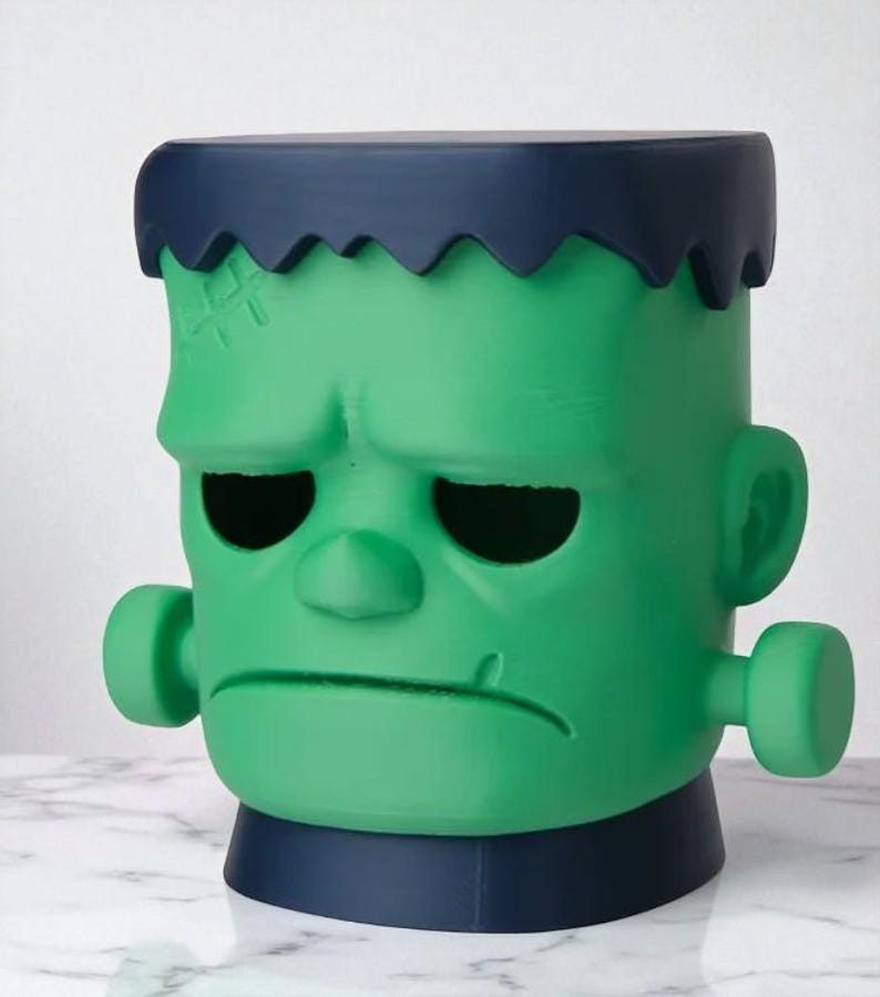 May include: A green Frankenstein-themed decorative container with a dark blue top and base. The container has a detailed face with stitched scars, black eye sockets, and side bolts. A unique Halloween or novelty item.