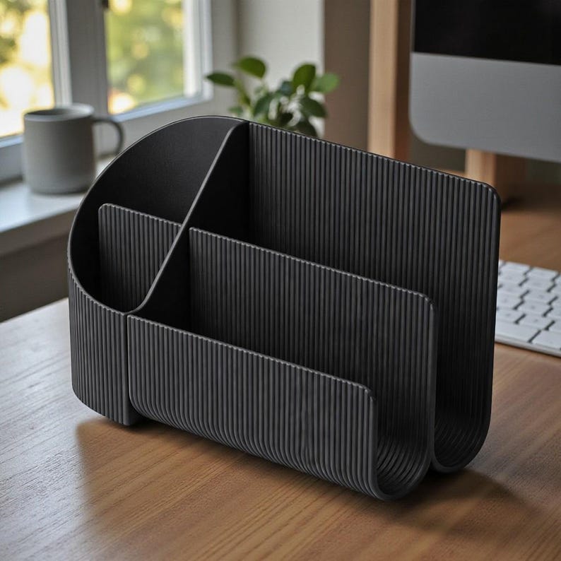 May include: A black desk organizer with multiple compartments. The organizer has a curved design with vertical ribbed detailing. It sits on a wooden desk near a window and a computer.