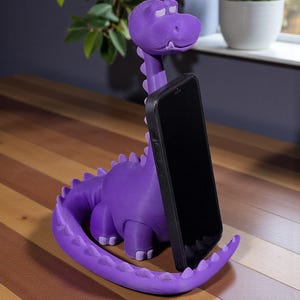 May include: A purple dinosaur-shaped phone holder with a black phone in place. The holder is made of a 3D-printed material and has a long neck, a tail, and small feet. The phone is held upright, and the holder is on a wooden surface.