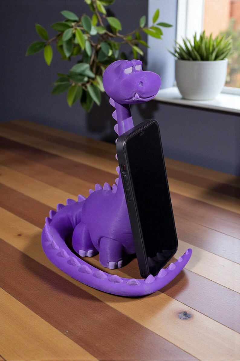May include: A purple dinosaur-shaped phone holder with a black phone in place. The holder is made of a 3D-printed material and has a long neck, a tail, and small feet. The phone is held upright, and the holder is on a wooden surface.