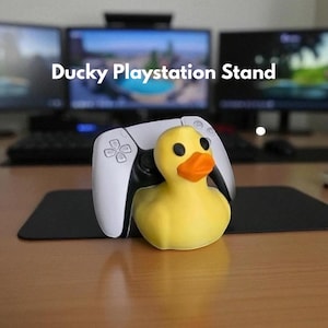 May include: A yellow rubber duck with a white Playstation controller. The duck has black eyes and an orange beak. The text "Ducky Playstation Stand" is displayed above the duck and controller. The background includes computer monitors and a desk.