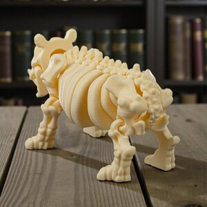 May include: A cream-colored, 3D-printed model of a skeletal animal, possibly a dog or similar quadruped. The model is detailed, showing ribs, spine, and leg bones. It stands on a wooden surface, with a blurred bookshelf background.