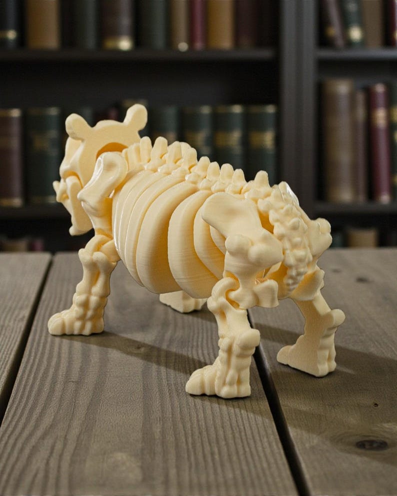May include: A cream-colored, 3D-printed model of a skeletal animal, possibly a dog or similar quadruped. The model is detailed, showing ribs, spine, and leg bones. It stands on a wooden surface, with a blurred bookshelf background.