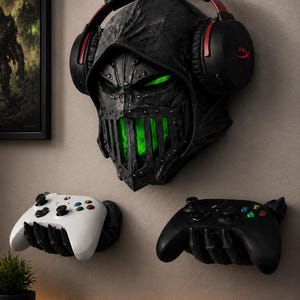 Könnte beinhalten: Eine an der Wand montierte schwarze Totenkopfmaske mit Kapuzendesign, leuchtend grünen Augen und Mund, die einen schwarz-roten Gaming-Kopfhörer trägt. Darunter halten zwei schwarze, klauenförmige Wandhalterungen einen weißen und einen schwarzen Controller.