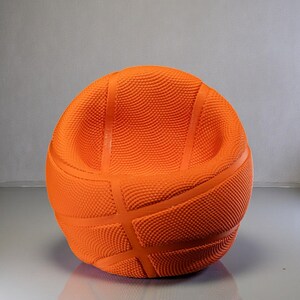 May include: An orange basketball-shaped chair with a textured surface and dark orange lines. The chair has a rounded shape with a low back and armrests, designed to resemble a basketball.