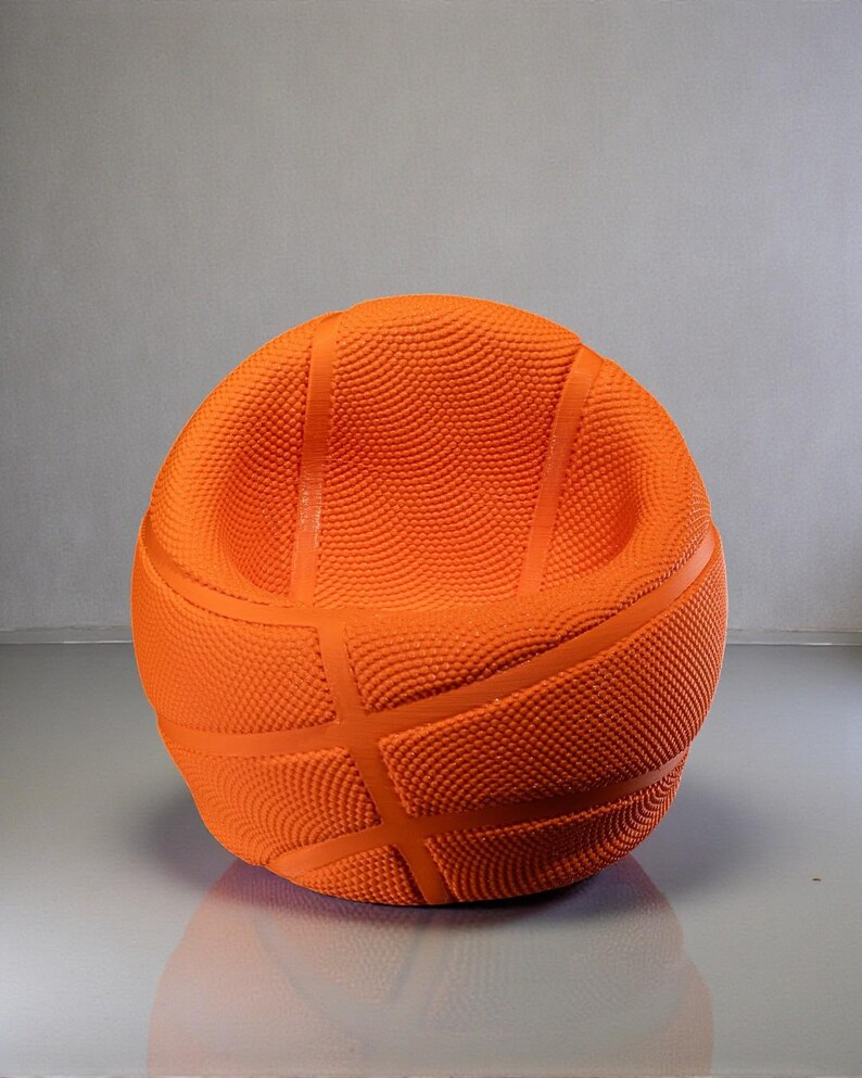 May include: An orange basketball-shaped chair with a textured surface and dark orange lines. The chair has a rounded shape with a low back and armrests, designed to resemble a basketball.