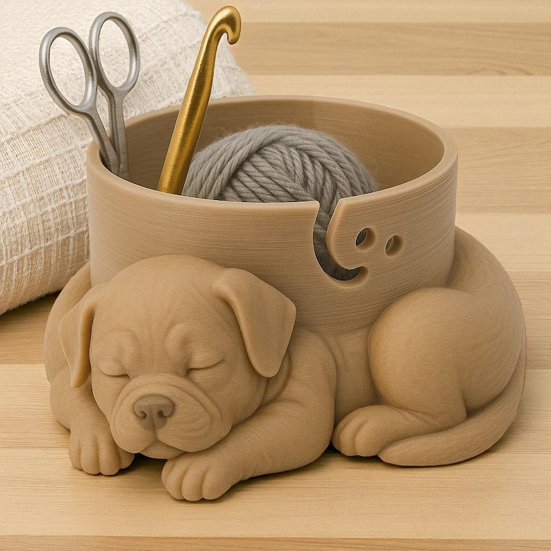 Puede incluir: Un cuenco de hilo de color beige con un dise&ntilde;o de cachorro durmiendo. El cuenco contiene una madeja de lana gris, un ganchillo dorado y unas tijeras plateadas. El cuenco tiene un agujero para que pase el hilo. El cachorro est&aacute; tumbado con los ojos cerrados.