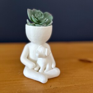 Puede incluir: Una figura de cer&aacute;mica blanca de una persona sentada con un perro, sosteniendo una planta suculenta en lugar de una cabeza. La figura est&aacute; sobre una superficie de madera con un fondo azul oscuro.