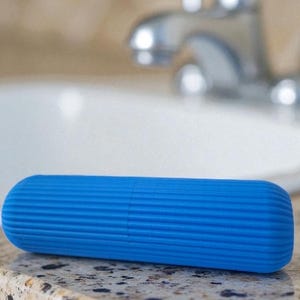 Pode incluir: Um estojo de escova de dentes azul brilhante, de formato cil&iacute;ndrico e com uma superf&iacute;cie canelada. O estojo &eacute; feito de um material s&oacute;lido e tem extremidades arredondadas. Descansa sobre uma bancada salpicada.
