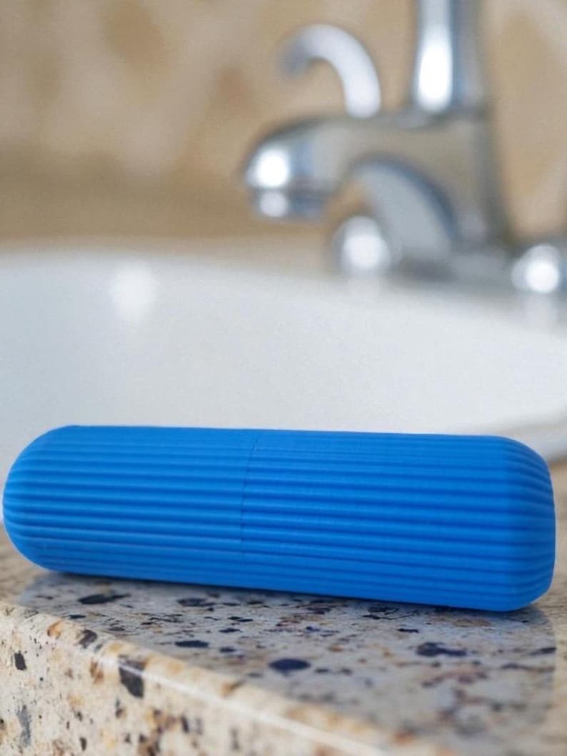 Pode incluir: Um estojo de escova de dentes azul brilhante, de formato cil&iacute;ndrico e com uma superf&iacute;cie canelada. O estojo &eacute; feito de um material s&oacute;lido e tem extremidades arredondadas. Descansa sobre uma bancada salpicada.