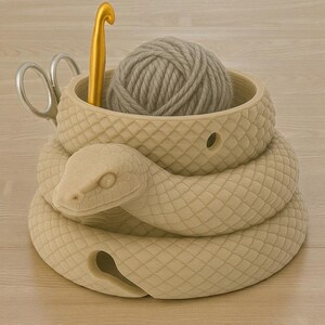 May include: A beige snake-shaped yarn bowl with a ball of gray yarn inside. A gold crochet hook and silver scissors are also present. The bowl has a textured, scale-like pattern. The snake's head is at the front.