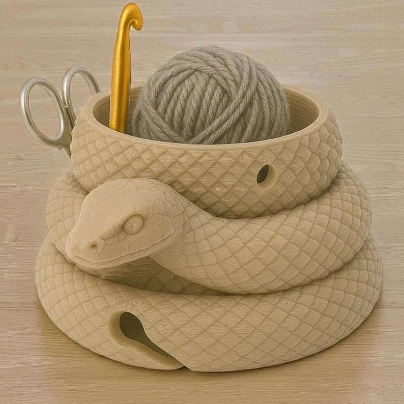 May include: A beige snake-shaped yarn bowl with a ball of gray yarn inside. A gold crochet hook and silver scissors are also present. The bowl has a textured, scale-like pattern. The snake's head is at the front.