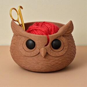 May include: A brown owl-shaped yarn bowl with black eyes and a red yarn ball inside. Gold scissors are placed in the bowl. The bowl has a textured surface and is designed to hold yarn while knitting or crocheting.