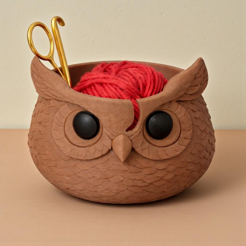 May include: A brown owl-shaped yarn bowl with black eyes and a red yarn ball inside. Gold scissors are placed in the bowl. The bowl has a textured surface and is designed to hold yarn while knitting or crocheting.
