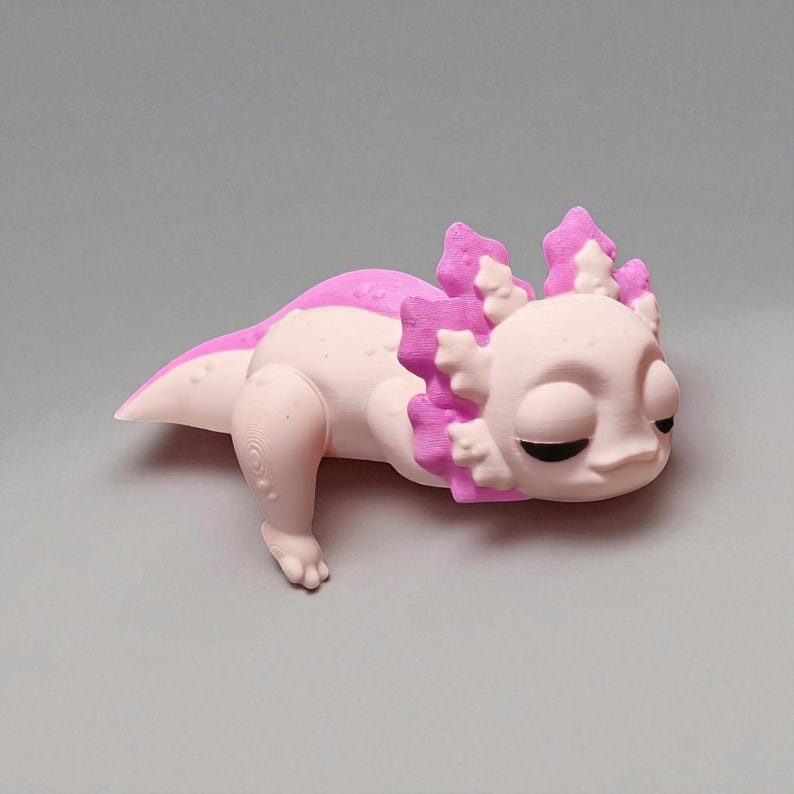 May include: A 3D-printed axolotl figurine in light pink and magenta. The salamander-like creature is lying down, with detailed gills and a rounded body. The axolotl has closed eyes and a smooth, matte finish.