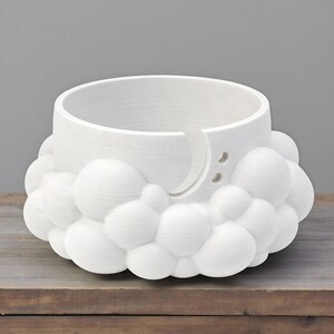 K&ouml;nnte beinhalten: Eine wei&szlig;e Garnschale mit wolkenf&ouml;rmigem Sockel und halbmondf&ouml;rmiger Garnf&uuml;hrung. Die Schale hat eine zylindrische Form mit zwei kleinen L&ouml;chern und einem gebogenen Schlitz f&uuml;r das Garn. Die Schale besteht aus einem wei&szlig;en Material.