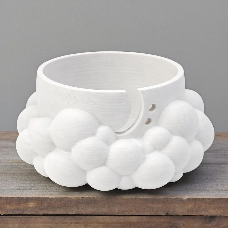 K&ouml;nnte beinhalten: Eine wei&szlig;e Garnschale mit wolkenf&ouml;rmigem Sockel und halbmondf&ouml;rmiger Garnf&uuml;hrung. Die Schale hat eine zylindrische Form mit zwei kleinen L&ouml;chern und einem gebogenen Schlitz f&uuml;r das Garn. Die Schale besteht aus einem wei&szlig;en Material.