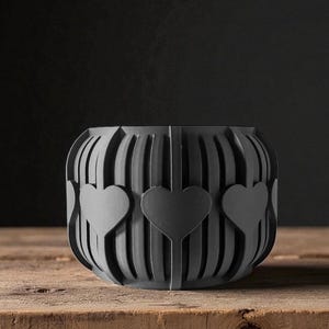 May include: A dark gray, heart-shaped decorative bowl. The bowl has a series of vertical lines with three heart cutouts. The bowl is sitting on a wooden surface.