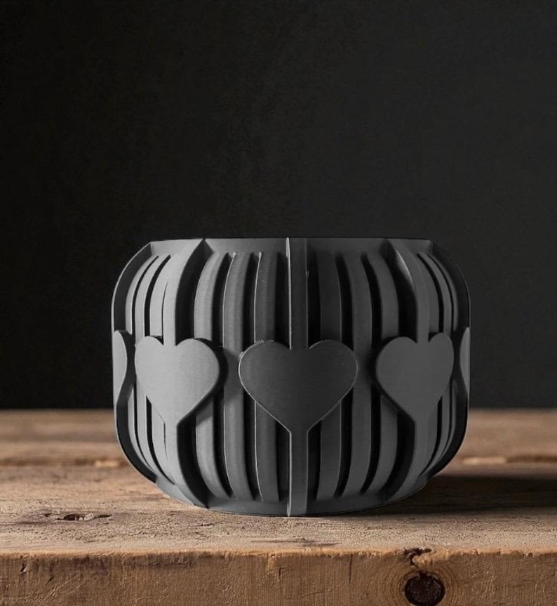 May include: A dark gray, heart-shaped decorative bowl. The bowl has a series of vertical lines with three heart cutouts. The bowl is sitting on a wooden surface.