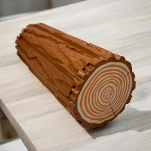 May include: A brown log-shaped object with a detailed wood grain texture. The end of the log features concentric circles in shades of orange and white, resembling tree rings. The object is designed to look like a realistic log.