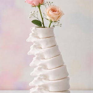 May include: A white ceramic vase shaped like a spine, holding two roses. One rose is light pink, the other is peach. The background is a soft gradient of pink, purple, and white.