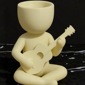 May include: A cream-colored figurine of a person seated cross-legged, playing a small guitar. The figure has a large, open head, suggesting a planter or container. The guitar has a round sound hole. The figurine is made of a smooth material.
