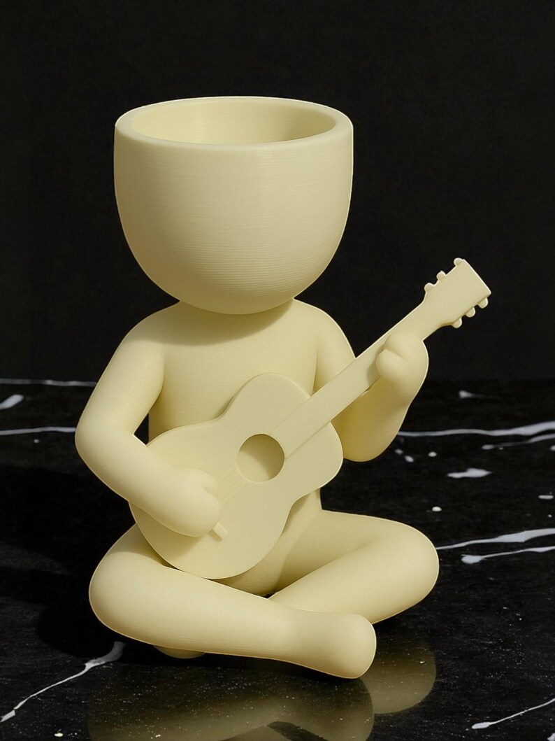 May include: A cream-colored figurine of a person seated cross-legged, playing a small guitar. The figure has a large, open head, suggesting a planter or container. The guitar has a round sound hole. The figurine is made of a smooth material.