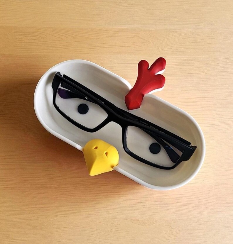 May include: A white ceramic tray holds a pair of black-framed eyeglasses, a red decorative piece, and a yellow heart-shaped object. The tray sits on a light wood surface. The eyeglasses have dark lenses.
