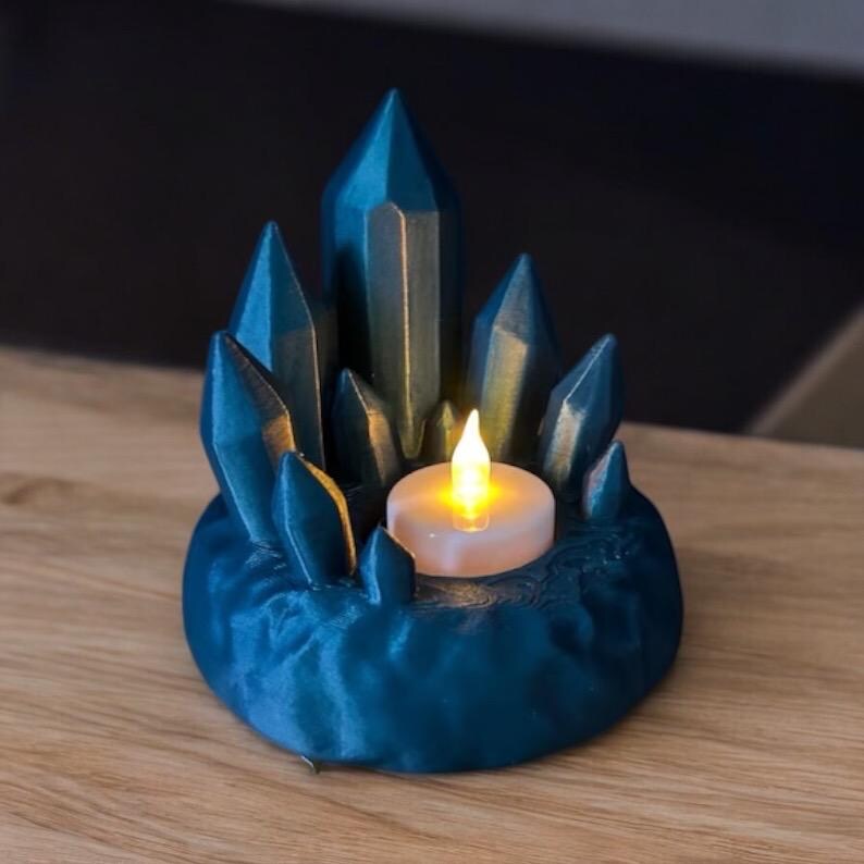 May include: A teal-colored crystal-shaped candle holder with a small, lit, battery-operated candle. The holder has a round base and several crystal-like structures surrounding the candle. The candle emits a warm, yellow glow.