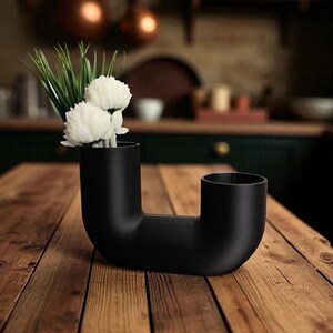 May include: A black, modern, sculptural vase with a unique, curved design. The vase holds white flowers and green grass, creating a striking contrast against the matte black finish. The vase is placed on a wooden table.