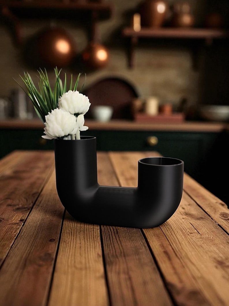 May include: A black, modern, sculptural vase with a unique, curved design. The vase holds white flowers and green grass, creating a striking contrast against the matte black finish. The vase is placed on a wooden table.
