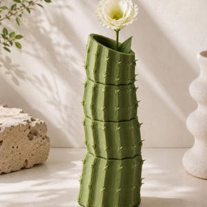 May include: A green cactus-shaped vase with a white flower. The vase is made of stacked sections, each with vertical ridges and small, pointed details. The flower has a large, ruffled bloom.