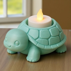 Peut inclure: Un bougeoir en forme de tortue turquoise avec un visage souriant et une carapace texturée. Le bougeoir contient une bougie blanche avec une flamme LED jaune vacillante. L'objet décoratif est en matériau solide et repose sur une surface en bois.