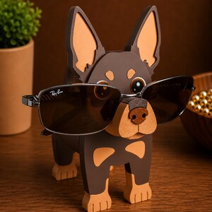 May include: A brown and tan dog-shaped eyeglass holder with a pair of Ray-Ban sunglasses. The holder has large ears and a textured nose. The sunglasses are dark-tinted with a silver frame.