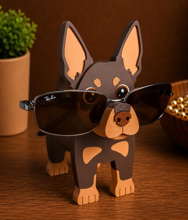 May include: A brown and tan dog-shaped eyeglass holder with a pair of Ray-Ban sunglasses. The holder has large ears and a textured nose. The sunglasses are dark-tinted with a silver frame.