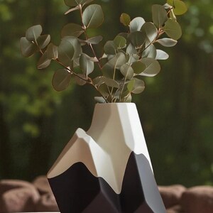 Op de afbeelding: Een moderne, geometrische vaas in de vorm van een bergketen. De vaas is voornamelijk zwart, wit en grijs, met een uniek hoekig ontwerp. Hij bevat een takje eucalyptus met groene bladeren, wat een natuurlijk contrast cre&euml;ert.