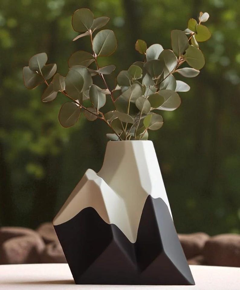 Op de afbeelding: Een moderne, geometrische vaas in de vorm van een bergketen. De vaas is voornamelijk zwart, wit en grijs, met een uniek hoekig ontwerp. Hij bevat een takje eucalyptus met groene bladeren, wat een natuurlijk contrast cre&euml;ert.