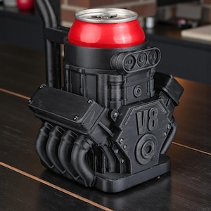 May include: A black V8 engine-shaped can holder with a red can. The holder has detailed features, including exhaust pipes and the text "V8". The can holder is on a dark wooden surface.