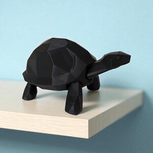 May include: A black geometric turtle figurine with a faceted design. The turtle is positioned on a light-colored shelf against a light blue background. The figurine has a modern, minimalist aesthetic.