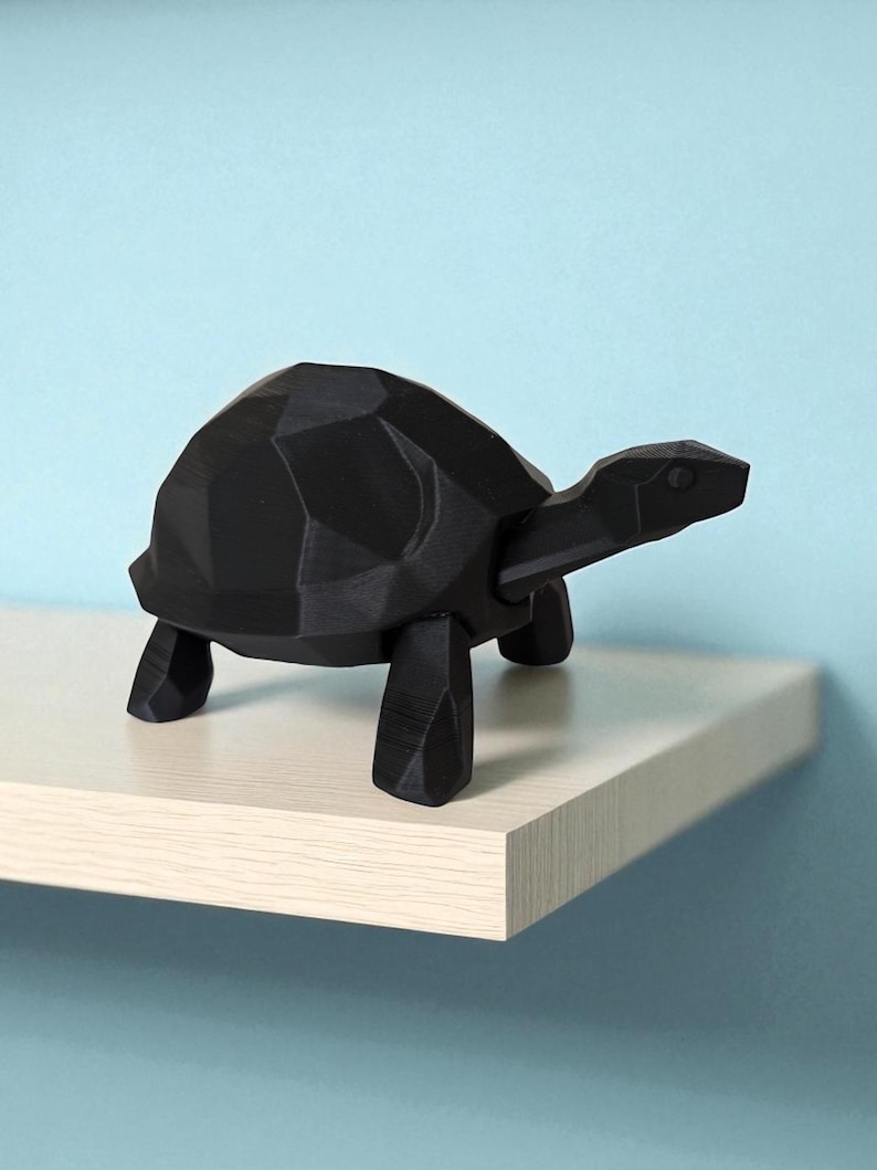 May include: A black geometric turtle figurine with a faceted design. The turtle is positioned on a light-colored shelf against a light blue background. The figurine has a modern, minimalist aesthetic.