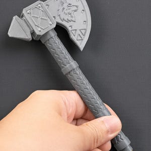 May include: A gray pen shaped like a battle axe. The axe head features a wolf design and geometric shapes. The handle has a textured pattern. The pen is held in a hand.
