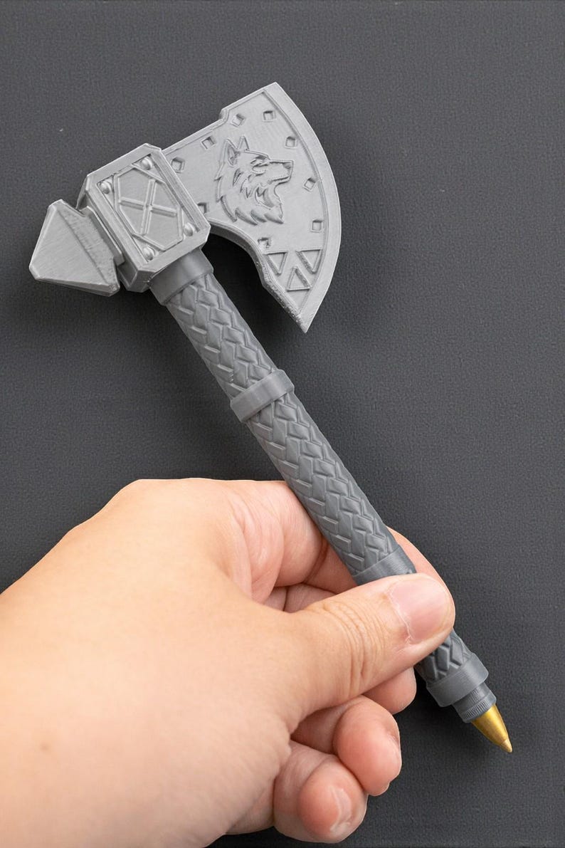 May include: A gray pen shaped like a battle axe. The axe head features a wolf design and geometric shapes. The handle has a textured pattern. The pen is held in a hand.