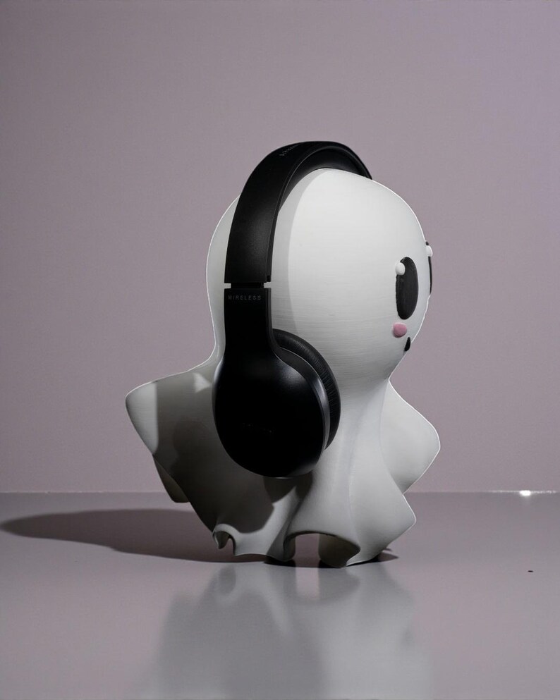 May include: A white ghost-shaped headphone stand with black headphones. The stand has a cartoonish design with black eyes and pink cheeks. The headphones are wireless and sit over the top of the ghost's head.